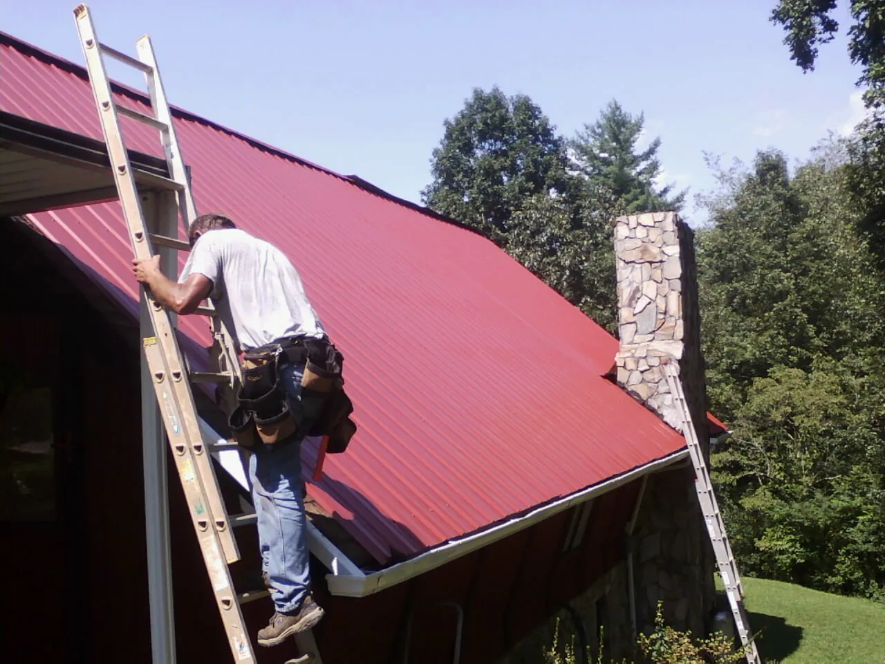 Roofer on a ladder beside a red metal roof and stone chimney — Soffit & Fascia Repair by Roofing Brothers in Summit